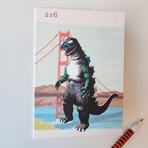 May include: A notebook featuring a Godzilla illustration in front of the Golden Gate Bridge. The dinosaur is black, green, and blue. The notebook has the number 216 in the upper left corner. A pen with orange and black stripes is in the lower right corner.