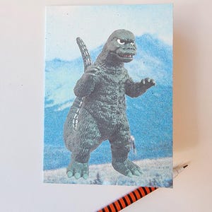 May include: A canvas print featuring a Godzilla figure against a backdrop of a mountain and blue sky. The Godzilla figure is gray and green. A black and orange striped pen is in the foreground.