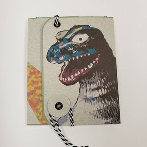 May include: A small, handmade book with a brown paper cover. The cover features a colorful illustration of a monster with a blue face and red mouth. The book is tied together with a black and white string.