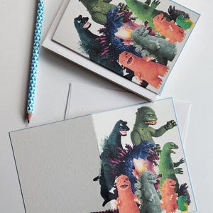 May include: Greeting cards featuring a colorful illustration of various dinosaur-like creatures. The cards have a light blue border and are paired with a blue and white patterned pencil with a yellow eraser.