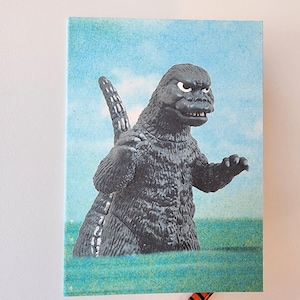May include: A vintage-style notebook featuring a Godzilla illustration. The image shows the iconic monster emerging from water, set against a blue sky background. The notebook has a retro aesthetic, with a pencil peeking out from the bottom.