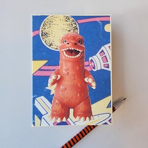 May include: A vintage-style toy dinosaur figurine in orange, set against a backdrop of a blue sky with a yellow moon and a space station. A black and orange striped pen rests nearby.