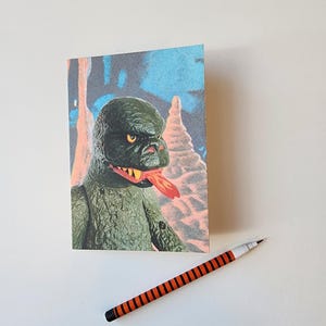 May include: A greeting card featuring a green monster with a red tongue, set against a blue and white background. A black and orange striped pen rests beside the card. The monster has yellow eyes and a textured appearance.