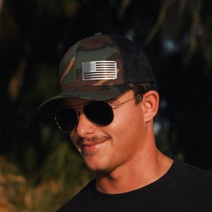 May include: A man wearing a camouflage and black trucker hat with a silver American flag patch. He is also wearing sunglasses and a black shirt.