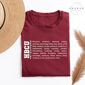 May include: Maroon t-shirt with the text "HBCU HOMECOMING" in white, along with a list of related words. The shirt is folded and placed on a light-colored surface, with a straw hat partially visible in the upper left corner.