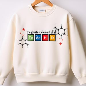 May include: Cream-colored sweatshirt with the text "the greatest element of all" above the word "TEACHER" spelled out using periodic table elements. The design includes red stars and molecular structures.
