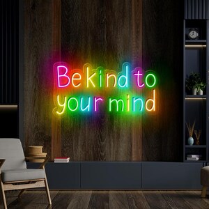 May include: Neon sign with rainbow colored letters that reads "Be kind to your mind". The sign is mounted on a dark wood wall.