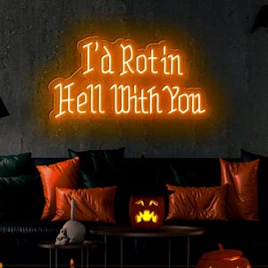 May include: An orange neon sign displaying the text "I'd Rot in Hell With You". Below, a black sofa is adorned with orange and green cushions. A skull candle holder and a carved pumpkin are also visible.