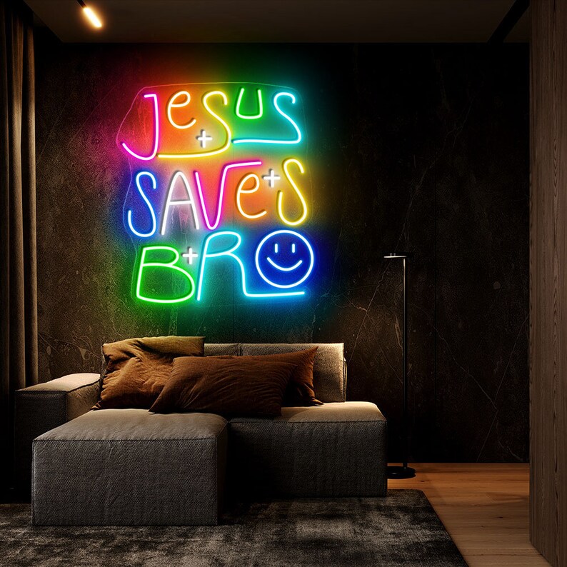 May include: A neon sign with the text "Jesus Saves Bro" in rainbow colors with a smiley face at the end. The sign is hanging on a dark wall above a gray couch.