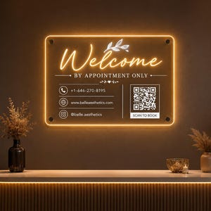 May include: A rectangular acrylic sign with a glowing orange LED border. The text reads Welcome BY APPOINTMENT ONLY, followed by a phone icon with 1-646-270-8195, a website icon with www.belleaesthetics.com, an Instagram icon with @belle.aesthetics, and a QR code.