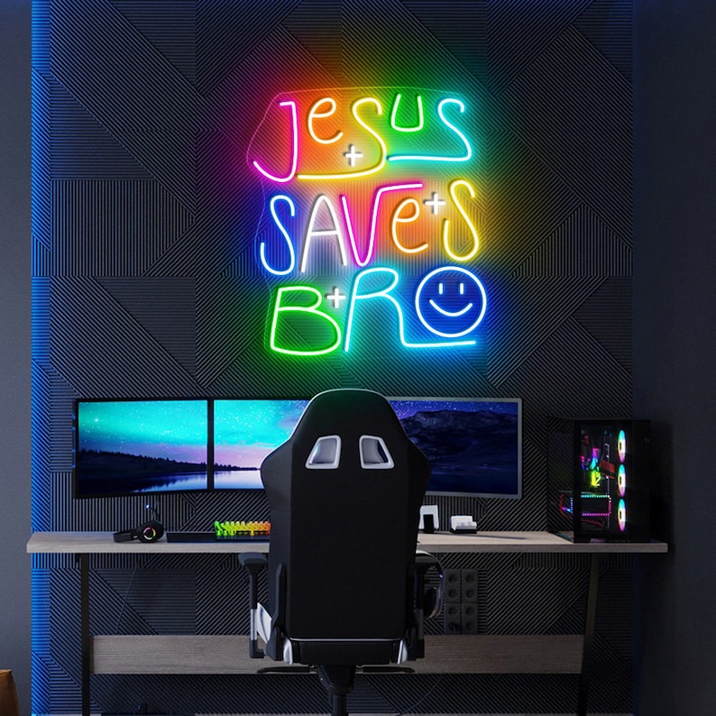 May include: A neon sign with the text "Jesus Saves Bro" in rainbow colors. The sign is hanging on a black wall with a gaming setup in front of it.