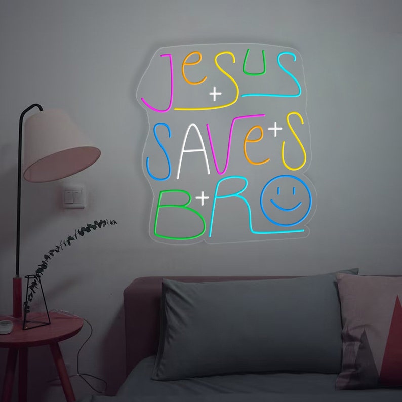 May include: Neon sign with the text "Jesus Saves Bro" in a colorful font with a smiley face. The sign is mounted on a white wall above a couch.