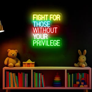 以下が含まれることがあります： 「FIGHT FOR THOSE WITHOUT YOUR PRIVILEGE」と書かれたネオンサイン。黄色、赤、緑色で表示されています。ピンク色の棚には本、テディベア、ウサギのぬいぐるみがあります。