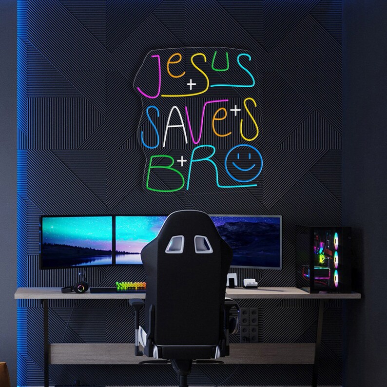 May include: A black gaming chair with white accents sits in front of a computer setup. A neon sign with the text "Jesus Saves Bro" in rainbow colors is on the wall behind the chair.