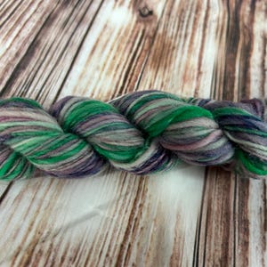 May include: A skein of hand-dyed yarn in shades of green, purple, and gray. The yarn is twisted and has a soft, textured appearance. The background is a wooden surface.