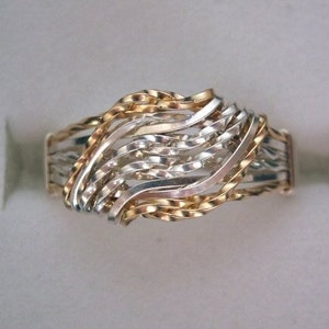 May include: A silver and gold ring with a woven design. The ring is made of multiple thin bands that are intertwined to create a wave-like pattern.