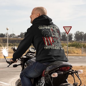 Può includere: Felpa con cappuccio nera con un design a bandiera americana invecchiata e la scritta "THE FIRST AMENDMENT DEFINES US, THE SECOND AMENDMENT DEFENDS US." Il design include fucili e stelle. La persona è in moto.