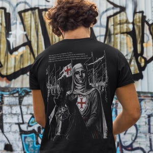 May include: Black t-shirt featuring a detailed illustration of a knight on horseback, with a red cross emblem. The design includes text at the top. The knight is depicted in a medieval style, with flags in the background. The shirt is a classic crew neck.