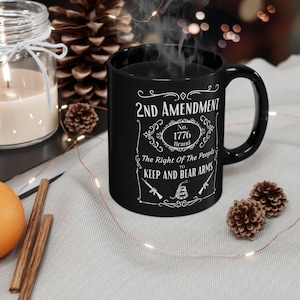 Pode incluir: Caneca de café preta com texto branco que diz "2nd Amendment No. 1776 Brand The Right Of The People To Keep And Bear Arms" com uma cobra e duas armas de fogo.