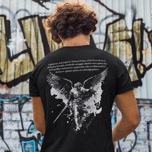Könnte beinhalten: Schwarzes T-Shirt mit einer detaillierten Grafik von Erzengel Michael mit Schwert und Flügeln. Das Design enthält Text aus einem Gebet. Das T-Shirt wird von einer Person vor einer Graffiti-Wand getragen.