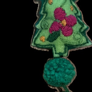 May include: A green felt Christmas tree bauble with a pink flower and gold accents. The bauble is attached to a green felt string with three green felt pom-poms.