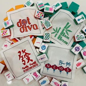 以下が含まれることがあります： 「dot diva」や「Bam Bitch」、漢字など、様々なデザインとテキストが刺繍された白いリネンのナプキン。緑、オレンジ、白の麻雀牌がナプキンを囲んでいます。