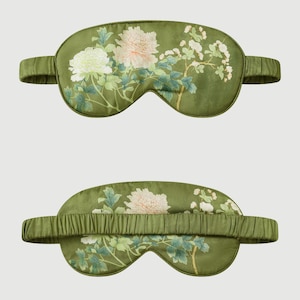 May include: Two olive green sleep masks with floral designs. The masks feature a pattern of white and pink flowers with green leaves and stems. The elastic band is the same olive green color as the mask.