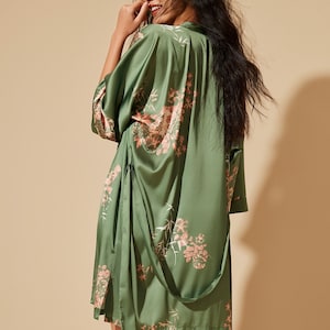 Puede incluir: Una bata de satén color verde salvia con un delicado estampado floral y de bambú en tonos rosa suave y blanco. La prenda presenta mangas anchas de tres cuartos, un diseño cruzado y un cinturón de tela a juego que cuelga suelto por el lado.