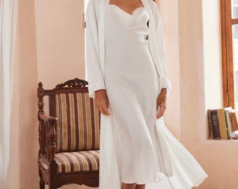 May include: A white satin dress with a cowl neckline and a matching long-sleeved robe. The dress falls to mid-calf length. The model is wearing light green slippers with feather trim. A wooden chair with striped upholstery is in the background.