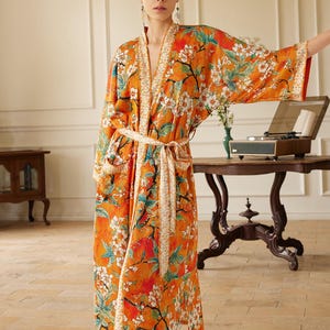 May include: An orange floral print robe with a cream-colored belt. The robe features a vibrant pattern of white flowers, green leaves, and red accents. The robe is made of a soft, flowing fabric and has wide sleeves.