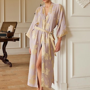 Peut inclure: Une robe lilas avec des motifs floraux et des bordures dorées. La robe est nouée à la taille avec une ceinture assortie. Les manches sont larges et la robe est ouverte sur le devant. La modèle porte des pantoufles assorties.