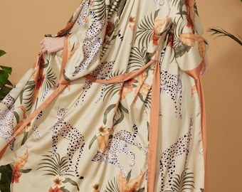 Puede incluir: Una larga bata de seda beige con estampado de guepardo y hojas tropicales. La bata tiene una faja naranja a juego.