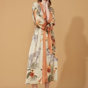 May include: A long, cream-colored robe with a vibrant floral and leopard print. The robe features a contrasting orange trim along the edges and a matching belt. The design includes tropical flowers and leopard spots.