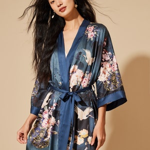 May include: A dark teal kimono-style robe with a floral and crane design. The robe has a navy blue trim and a matching belt. The floral pattern includes pink and white flowers. The robe is made of a silky material.