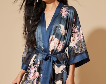 May include: A dark teal kimono-style robe with a floral and crane design. The robe has a navy blue trim and a matching belt. The floral pattern includes pink and white flowers. The robe is made of a silky material.