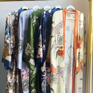 May include: A collection of silk robes in various colors and floral patterns hang on a gold clothing rack. The robes feature designs with flowers, leaves, and animal prints. The robes are in shades of blue, green, and cream.