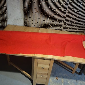 Peut inclure: Un long tissu rouge vif est drap&eacute; sur une table en bois clair. La table a un design &agrave; abattant et un petit meuble avec trois tiroirs. L'arri&egrave;re-plan pr&eacute;sente un motif g&eacute;om&eacute;trique noir et blanc.