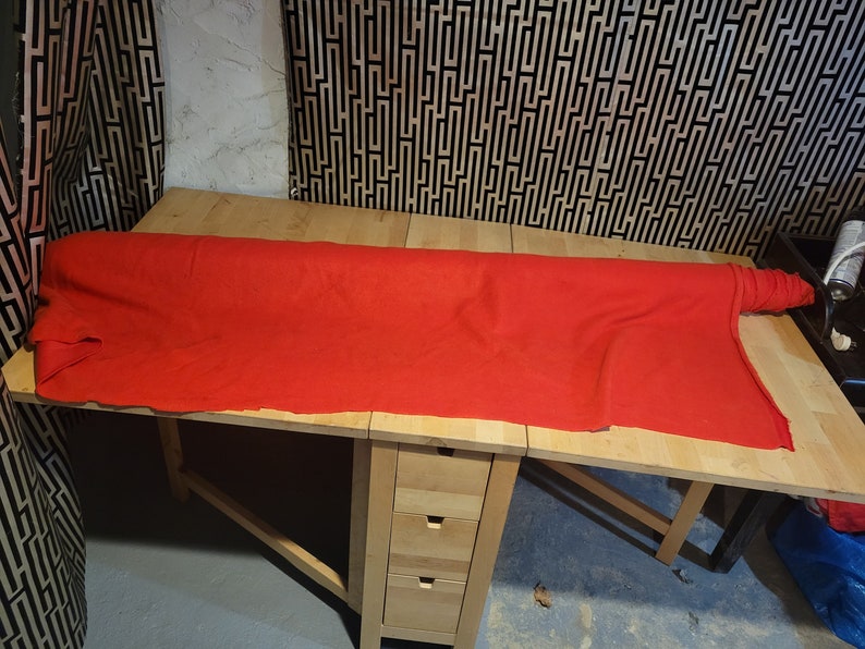 Peut inclure: Un long tissu rouge vif est drap&eacute; sur une table en bois clair. La table a un design &agrave; abattant et un petit meuble avec trois tiroirs. L'arri&egrave;re-plan pr&eacute;sente un motif g&eacute;om&eacute;trique noir et blanc.