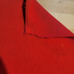 May include: A vibrant red fabric, possibly felt or a similar material, is partially folded over, revealing its textured surface. The edges show a raw, unfinished appearance. The fabric is a solid, bright red.
