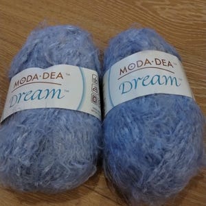Pode incluir: Duas meadas de lã azul clara com a marca "MODA-DEA" e a palavra "Dream" impressa na etiqueta. A lã tem uma textura macia e fofa, ideal para projetos de tricô ou crochê. A lã é de cor azul pastel lisa.