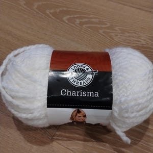 Puede incluir: Una gran madeja de lana blanca Charisma de Loops & Threads. La lana está envuelta con una etiqueta con el nombre de la marca y la imagen de una mujer. La lana está sobre una superficie de madera.