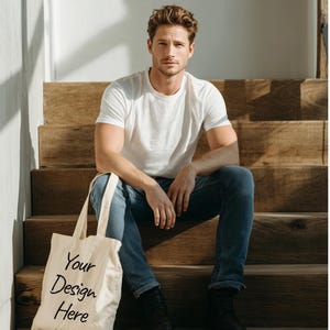Puede incluir: Una persona con una camiseta blanca y vaqueros azules está sentada en una escalera de madera, sosteniendo una bolsa de lona. La bolsa de lona tiene el texto "Your Design Here" en escritura negra. La persona lleva botas negras.