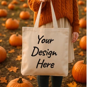 Puede incluir: Un bolso tote beige con el texto "Your Design Here" en negro. El bolso se sostiene frente a un fondo de calabazas y follaje otoñal. El bolso tote tiene asas largas.