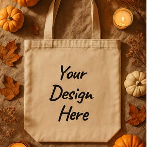 Puede incluir: Un bolso tote beige con el texto "Your Design Here" en negro. La bolsa está rodeada de calabazas, velas y hojas de otoño, creando un tema de temporada. La imagen tiene una estética cálida y acogedora.