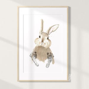 Könnte beinhalten: Aquarelldruck eines Hasen mit Fußabdrücken. Der Hase ist hellbraun mit rosa Akzenten in den Ohren. Die Fußabdrücke des Hasen sind grau. Der Druck befindet sich in einem hellen Holzrahmen.