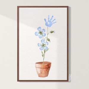 May include: A watercolor painting of a blue flower in a terracotta pot with a blue handprint above it. The painting is framed in a brown frame and signed "Mama & Charlie".
