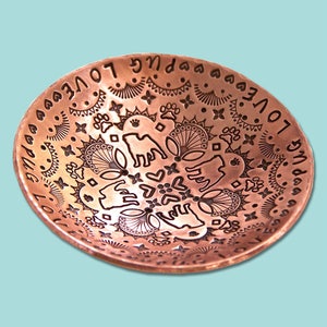 May include: A copper bowl with an intricate design engraved on the inside. The design features a repeating pattern of paw prints, hearts, and flowers. The words "Love Pug" are engraved around the rim of the bowl.