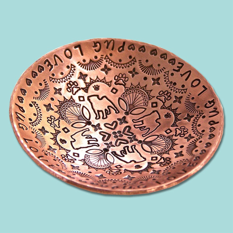 May include: A copper bowl with an intricate design engraved on the inside. The design features a repeating pattern of paw prints, hearts, and flowers. The words "Love Pug" are engraved around the rim of the bowl.