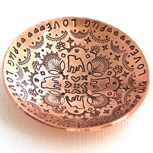 May include: A copper bowl with an intricate design featuring a pug dog, paw prints, hearts, and the words "Pug Love" repeated around the rim.