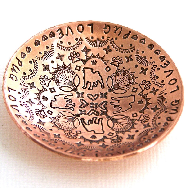 May include: A copper bowl with an intricate design featuring a pug dog, paw prints, hearts, and the words "Pug Love" repeated around the rim.